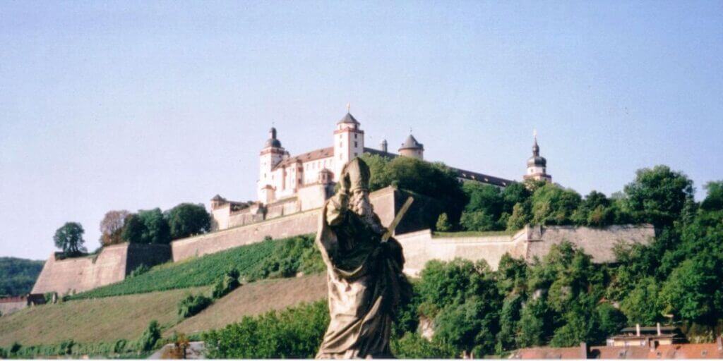 A picturesque view of Marienberg Fortress perched on a lush green hill in Wรผrzburg, Germany. The image foreground features a statue, adding depth and historical context, while the fortress itself is shown with its prominent towers and walls under a bright, clear sky.