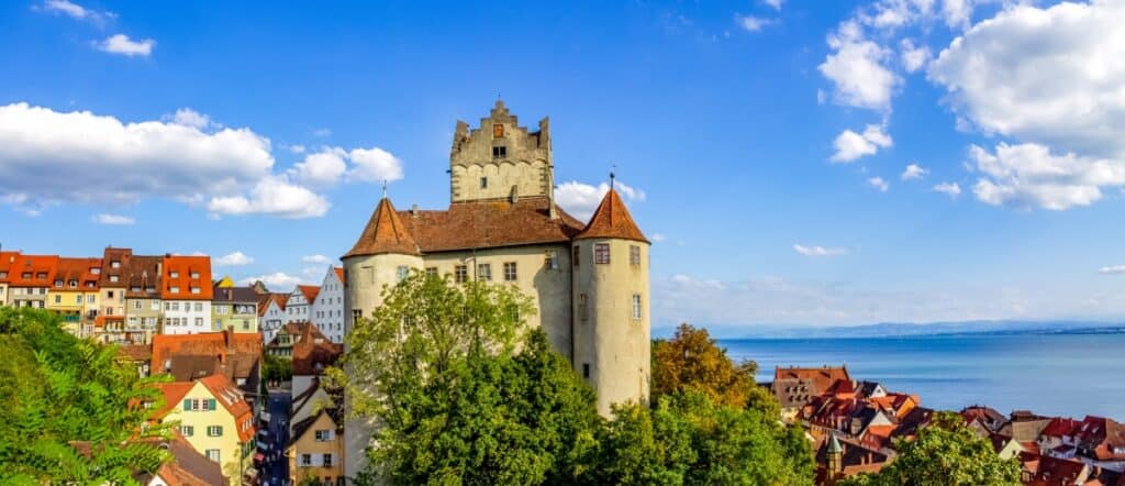 A panoramic view of Meersburg Castle, towering over the historic town by the shores of Lake Constance in Germany. The castle, with its distinctive white and reddish rooftops, stands majestically against a lush green backdrop and a clear blue sky. Below, the tightly packed, colorful houses of the town cascade towards the lake, presenting a charming old-world European scene.