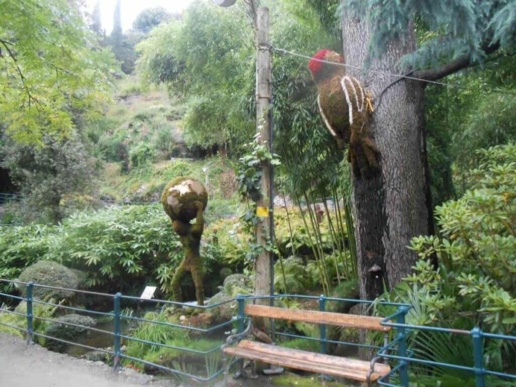This image captures a charming and whimsical scene at the Castel Trauttmansdorff Botanical Gardens in Merano, Italy. In the foreground, a whimsical sculpture resembling a golden frog stands atop a pole, overlooking a verdant garden with dense green foliage and various plant species. A rustic wooden bench sits by a metal railing