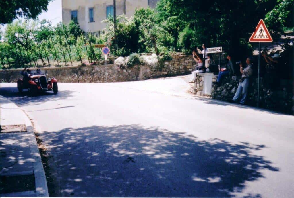 A vintage red sports car participating in the Mille Miglia rally takes a corner on a shaded Italian street, while spectators relax against a stone wall under the dappled sunlight filtered through trees.