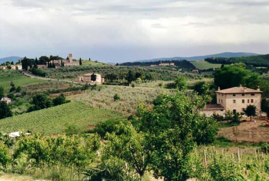 Scenic landscape of the countryside near Montepulciano, Tuscany, with a cluster of traditional Italian houses amidst lush vineyards, olive groves, and a historic tower in the distance under a cloudy sky.