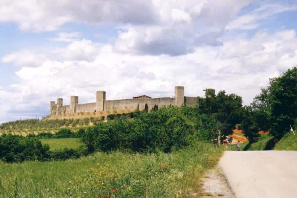 The imposing medieval walls of Monteriggioni, Tuscany, stand atop a hill surrounded by vibrant greenery. A gravel path leads towards the ancient fortress under a dynamic sky, showcasing the enduring legacy of Tuscany's historical architecture.