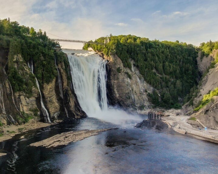 Montmorency Falls in Quebec, a powerful cascade pouring over steep cliffs surrounded by dense greenery. A suspension bridge spans the top of the falls, offering visitors a stunning view of this natural wonder and the mist rising from the plunging waters.