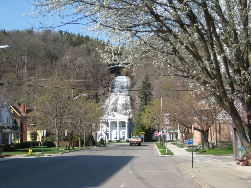 A unique view of Main Street in Montour Falls, New York, where a large waterfall cascades down a steep cliff directly at the end of the street. The street is lined with charming residential houses and budding spring trees, creating a picturesque small-town scene with the natural waterfall as a dramatic backdrop under a clear sky.