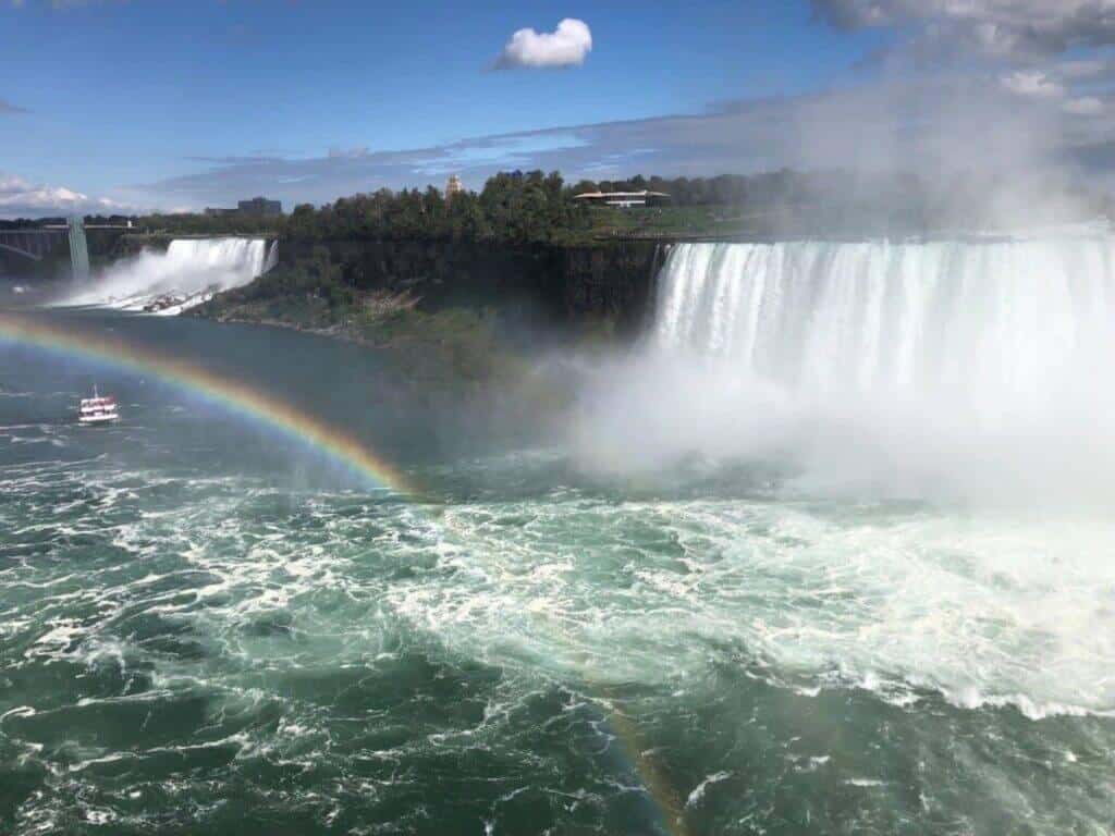 Breathtaking view of Niagara Falls, with a vivid rainbow arching across the misty air. The powerful falls pour vast amounts of water over the precipice, creating a dynamic scene of natural beauty. A tourist boat, visible near the base of the falls, appears small against the massive water flow. The scene is framed by a bright blue sky and lush greenery on the surrounding land