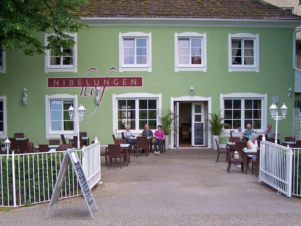 Patrons relax on the outdoor patio of the Nibelungen Hof Hotel in Tulln, with its pastel green facade and elegant white windows, inviting guests for a comfortable stay and pleasant dining experience