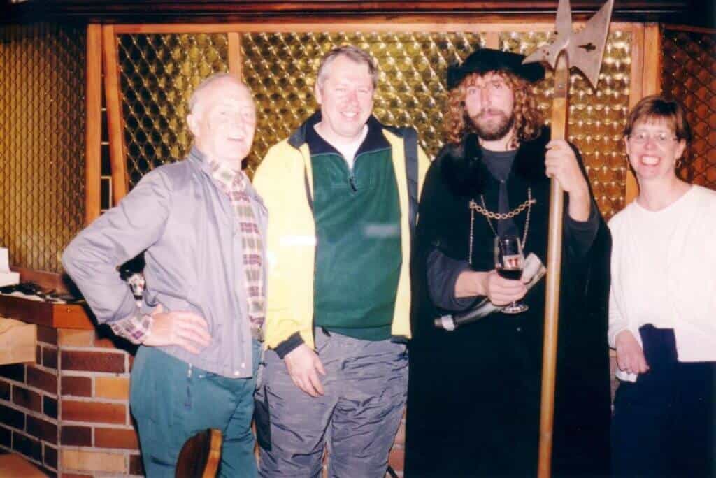 A group of tourists enjoying the Night Watchman tour in Rothenburg, Germany. The image shows four people, one dressed as a medieval night watchman complete with a cloak and halberd, posing together indoors. Their smiles and relaxed postures suggest a fun and engaging historical experience.