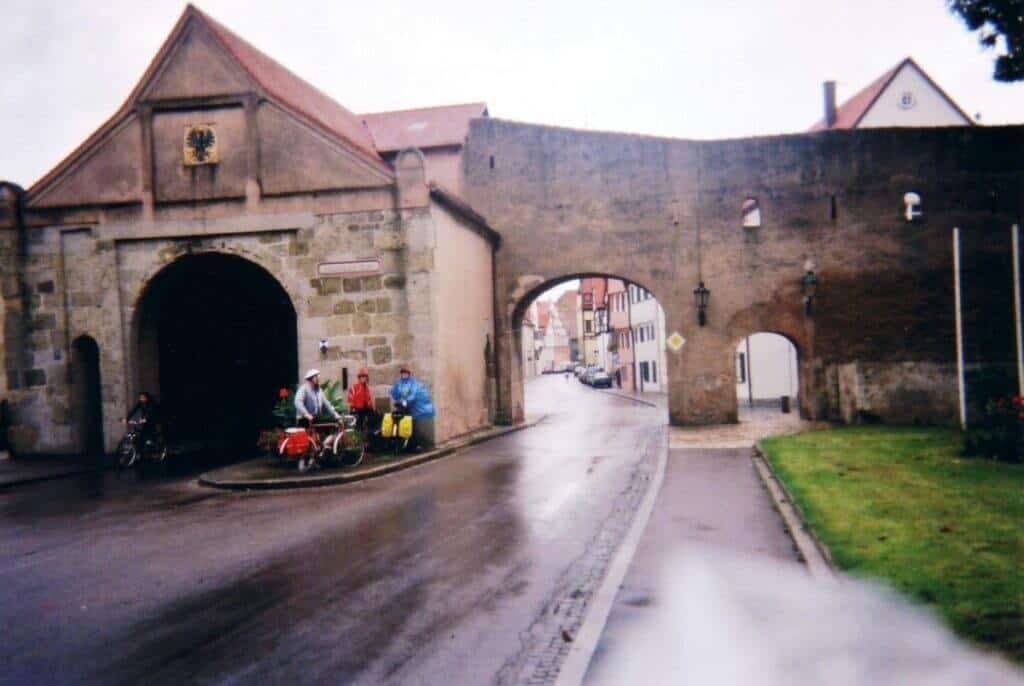 A group of cyclists in colorful rain gear pause under the historical stone archway of the Nรถrdlingen city walls in Germany. The wet cobblestone street reflects a cloudy day, while the ancient walls and arched gateways provide a glimpse into the town's medieval past.