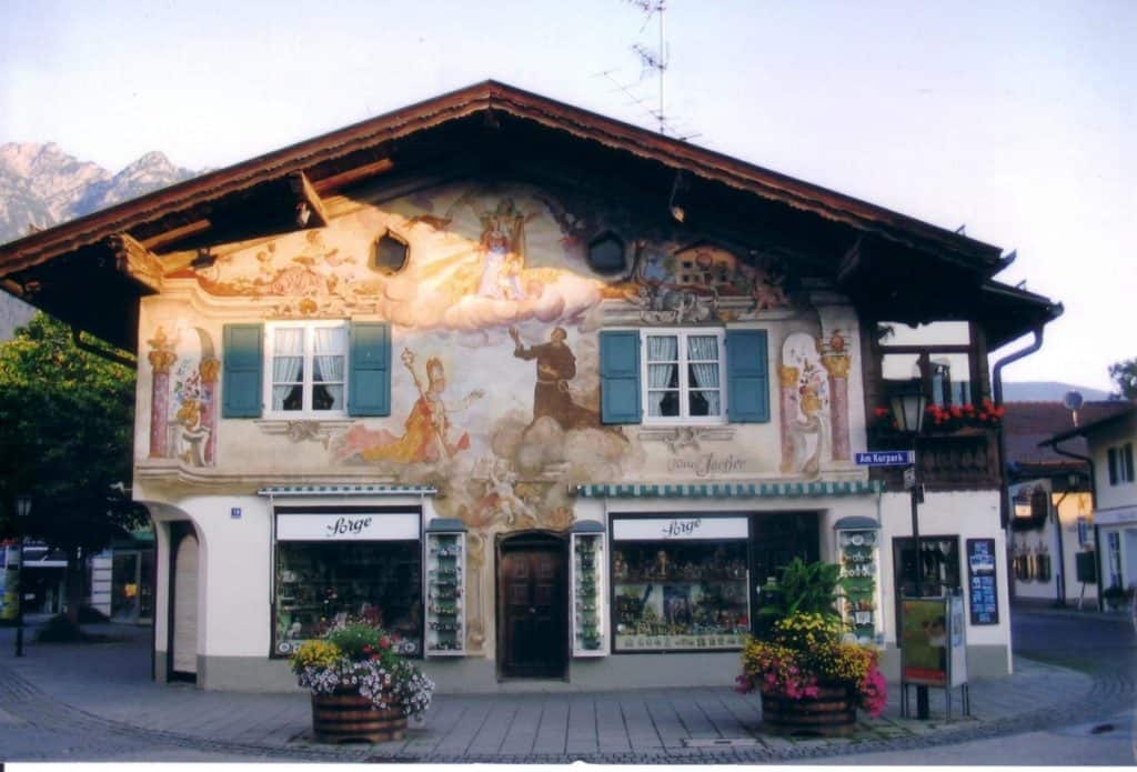 Traditional painted building in Garmisch-Partenkirchen, Germany, adorned with frescoes depicting folk scenes and floral motifs, flanked by windows with green shutters and vibrant flower boxes, set against a backdrop of alpine mountains.