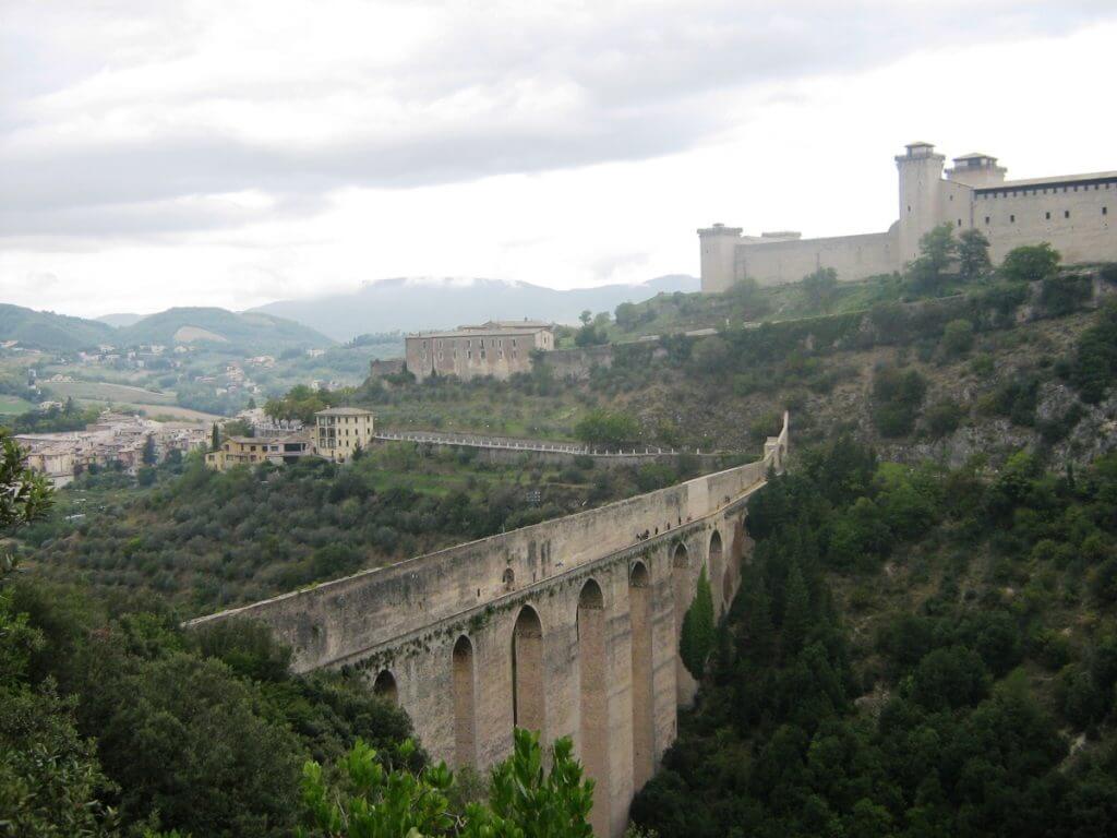 A dramatic view of the Ponte delle Torri, a medieval stone bridge, spanning a deep gorge with the Rocca Albornoziana fortress in the background, located in Spoleto, Italy. The bridge connects the fortress to the surrounding hillside, which is covered with lush greenery. The overcast sky adds a moody atmosphere to the historic and architectural marvel.