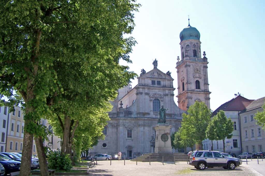 St. Stephen's Cathedral in Passau, Germany, stands prominently under a clear sky, framed by lush green trees. The cathedral features a grand facade with intricate architectural details and twin towers capped with green domes. In the foreground, a statue on a pedestal adds historical significance to the peaceful, sunlit square surrounded by parked cars and a few pedestrians