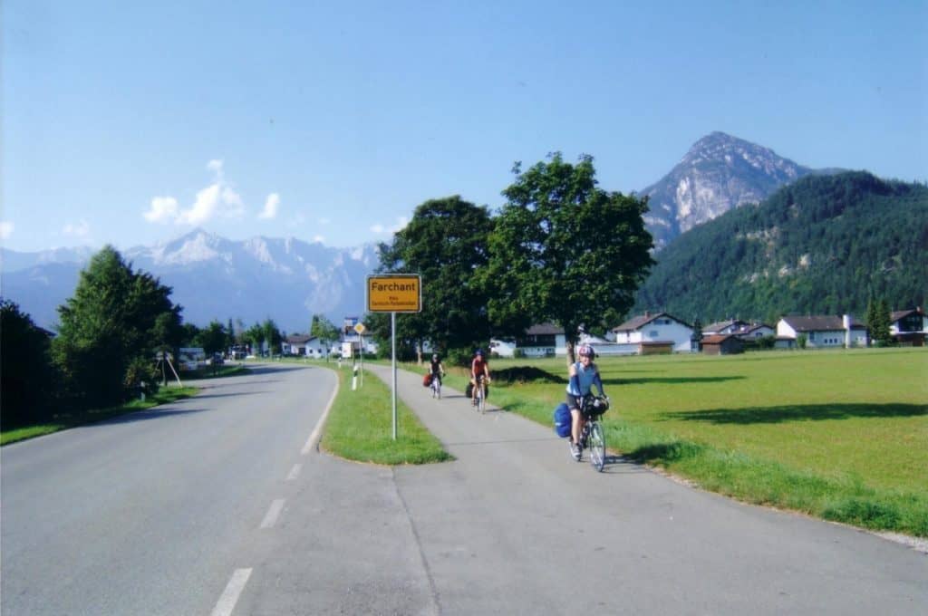 Bodensee to Konigssee Bike Route with cyclists on a sunny day departing Garmisch-Partenkirchen, Germany, with a road sign for Farchant in the foreground, a clear blue sky above, and the majestic Alps providing a breathtaking backdrop