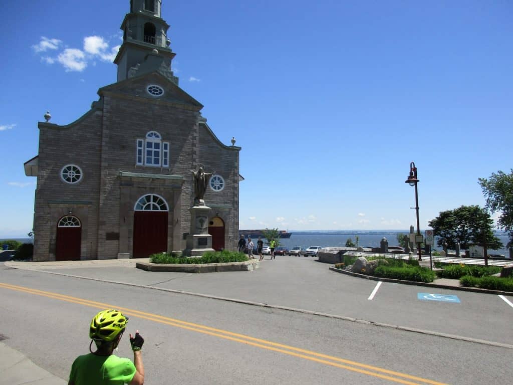 A cyclist in a bright green jersey gestures towards the historic church of Saint-Jean on Île d'Orléans, Quebec, with the Saint Lawrence River in the background. The clear blue sky and the open road create an inviting atmosphere