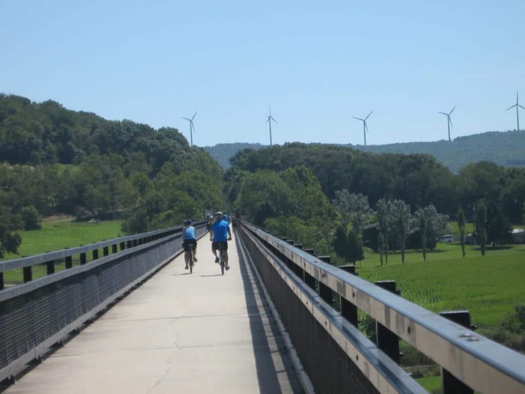 "Cyclists approach the Eastern Continental Divide tunnel on the Great Allegheny Passage Trail, framed by murals depicting local history and landscapes on its concrete walls under a sunny sky.