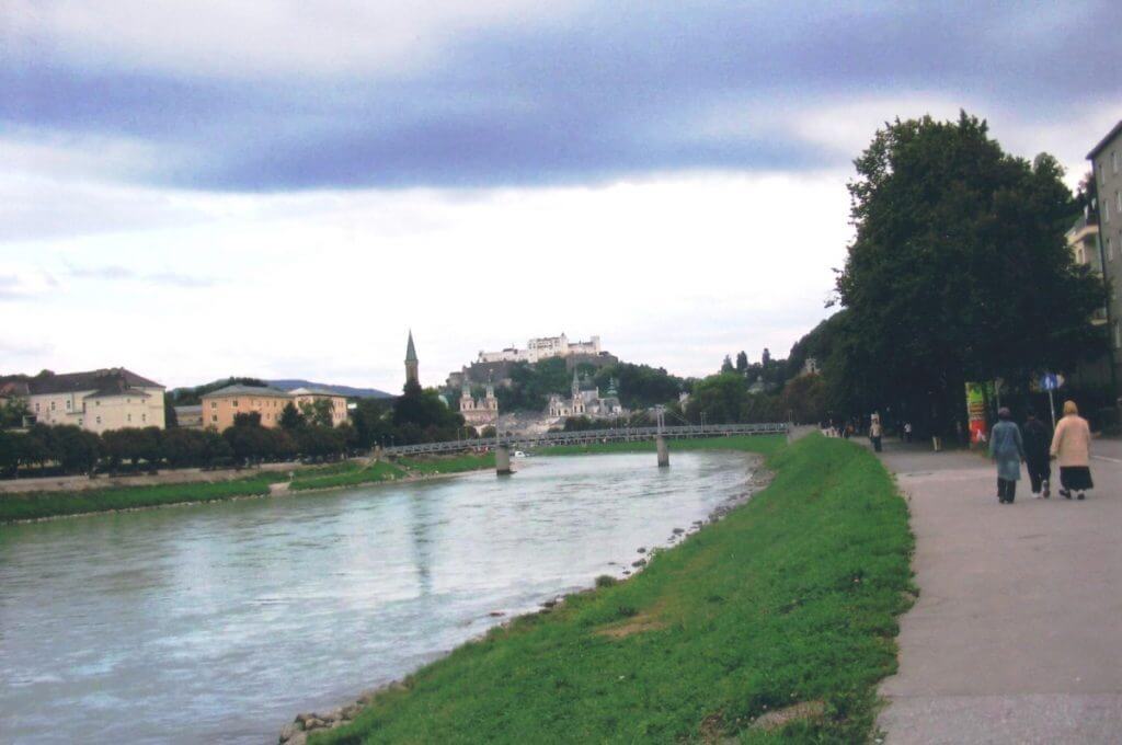 A peaceful riverside promenade in Salzburg, with people walking and pushing a shopping trolley along a pathway beside the gently flowing river. The landscape includes a lush green riverbank on one side and a line of historic buildings with distinctive European architecture on the other. In the background, the city's famous fortress perches atop a hill, partially obscured by clouds, adding a dramatic touch to the scenic urban view.