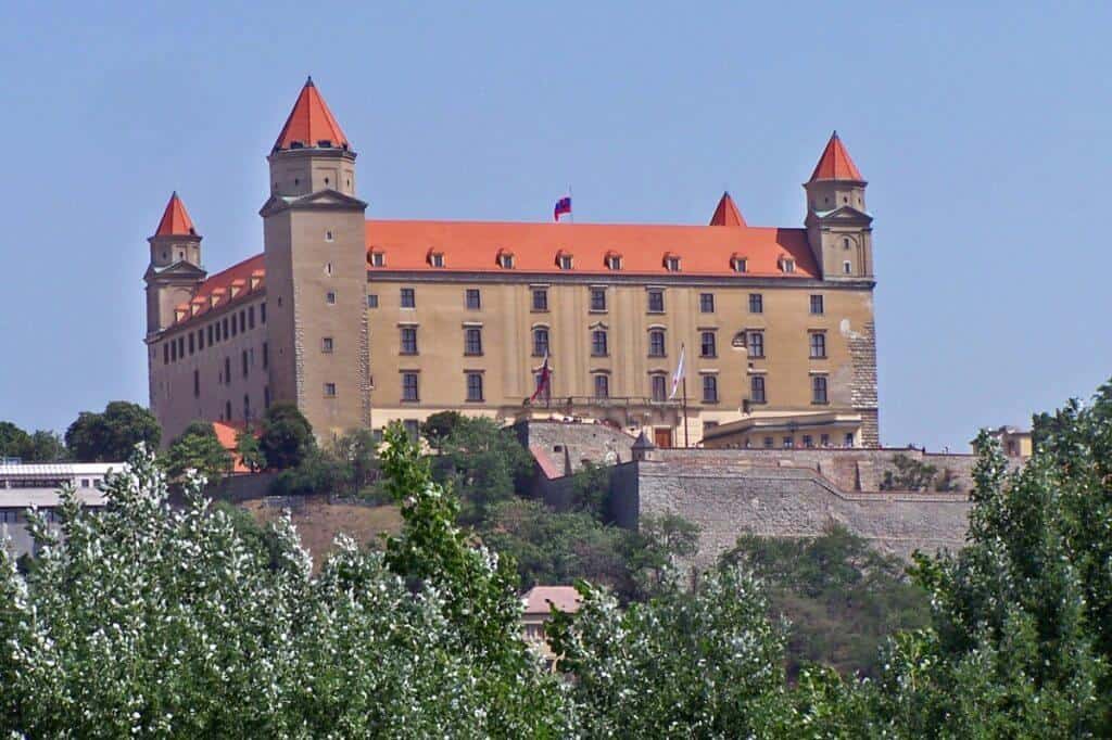 Bratislava Castle, a prominent historical landmark in Slovakia, stands majestically on a hill overlooking the city. The castle features a distinctive large rectangular building with four corner towers, each capped with a red roof, against a backdrop of lush green foliage and a clear blue sky.