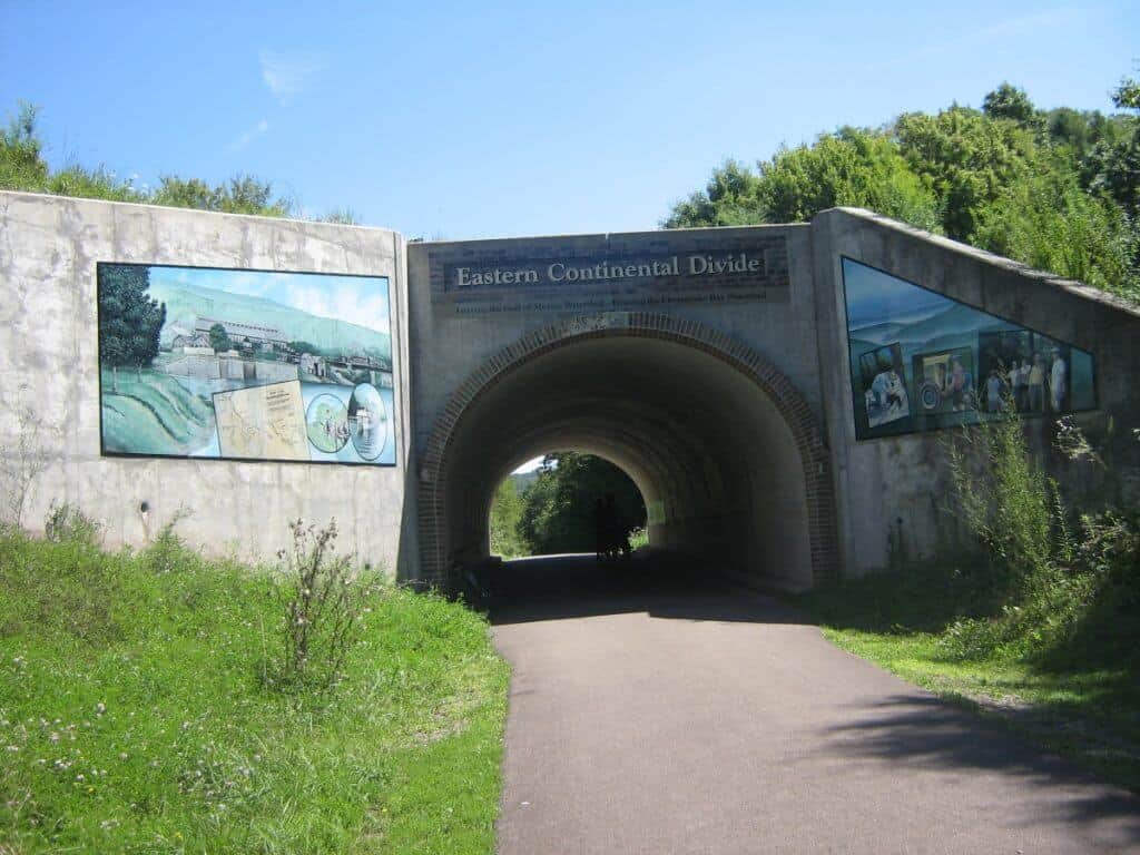 The Eastern Continental Divide landmark on the Great Allegheny Passage, featuring educational murals on either side of a tunnel entrance, under a bright blue sky.