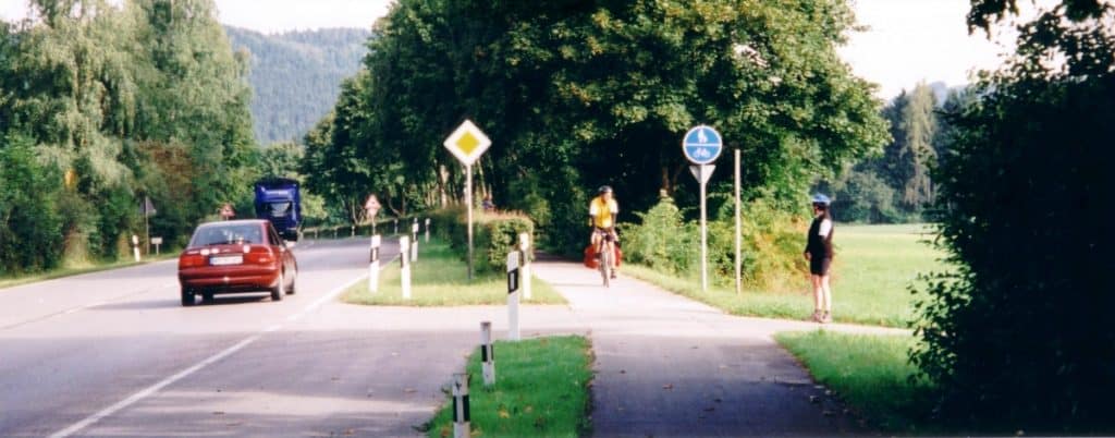 Cyclists and a pedestrian share a scenic path just outside Schongau, Germany. A paved cycling track runs parallel to the road, bordered by lush green trees, providing a peaceful route for outdoor activities. A car drives by, illustrating the coexistence of different modes of transport in this serene rural setting.