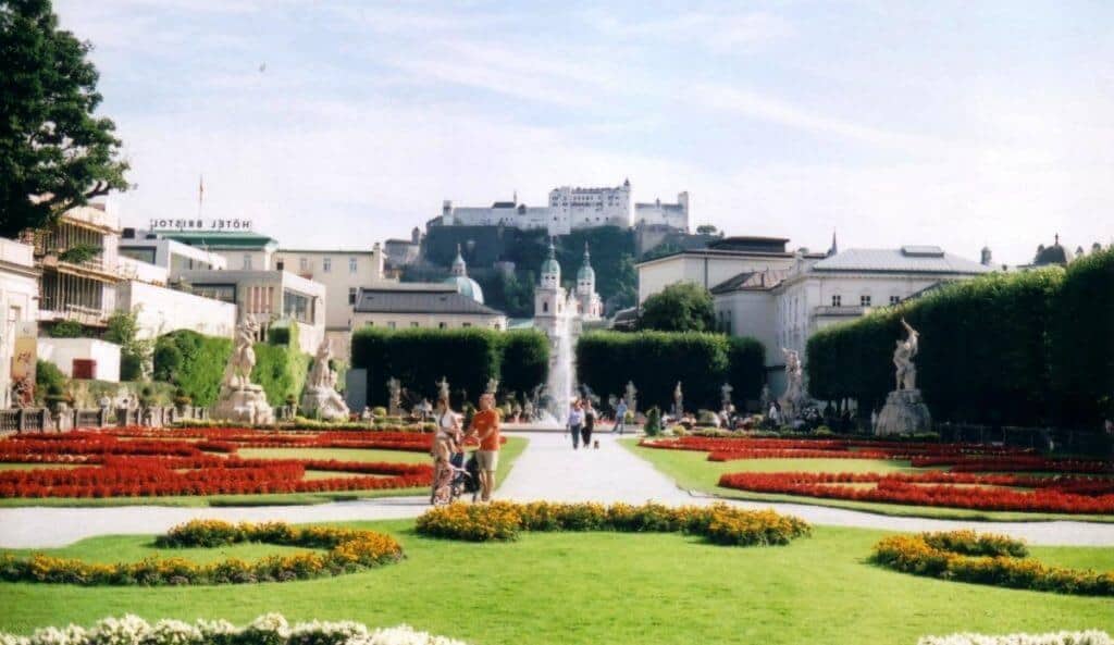 The image captures a vibrant view of Mirabell Gardens in Salzburg, Austria. In the foreground, beautifully manicured red and yellow flower beds and neatly trimmed hedges lead up to a central fountain surrounded by statues. In the background, the historic Hohensalzburg Fortress looms over the scene, providing a dramatic contrast to the serene garden in the sunshine. Two visitors can be seen near the fountain, enhancing the lively, tourist-friendly atmosphere of the gardens.