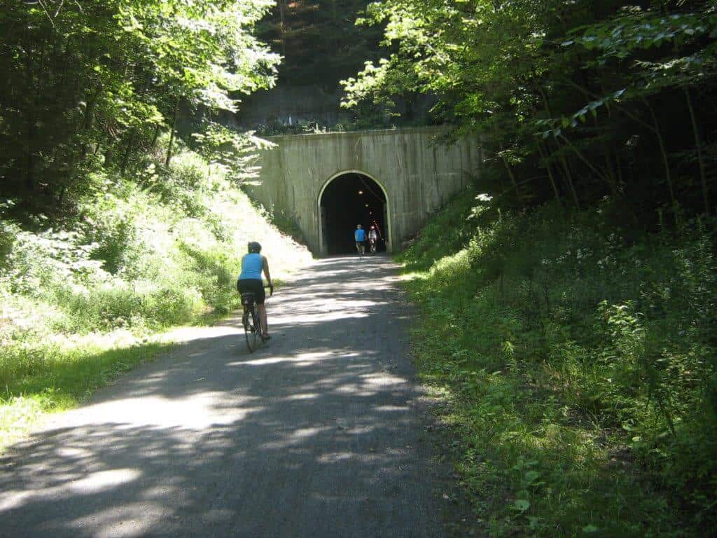 Cyclists ride towards the entrance of the Big Savage Tunnel, a well-known feature on the Great Allegheny Passage, surrounded by the lush greenery of a summer day.