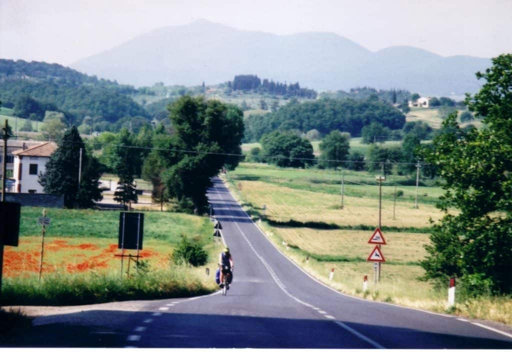 Bike Tours in Tuscany with a cyclist on a serene road near Chiusi, Tuscany, with a backdrop of gentle hills, lush greenery, and wildflowers. Road signs indicating a right turn and road narrows are visible, adding to the rural charm of the scene.