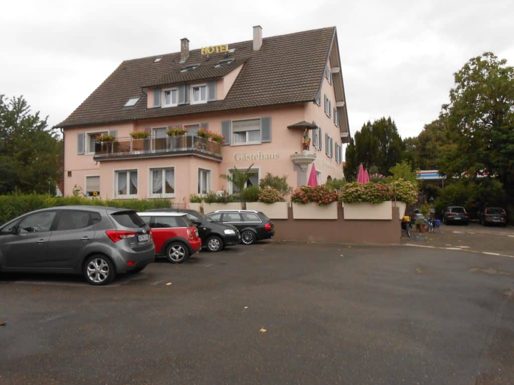 The Hotel Breisacherhof in Breisgau, Germany, presents a welcoming pink facade adorned with flower boxes, viewed from a parking area with several cars. The modest, traditional European architecture and the muted colors give a sense of the local hospitality and charm