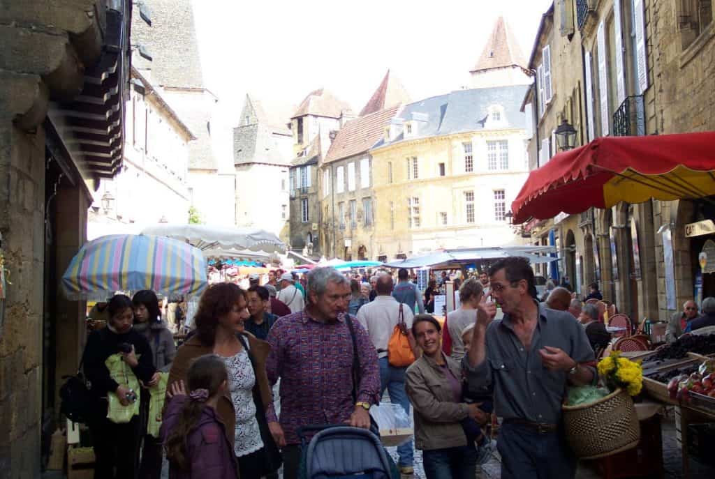 Vibrant atmosphere at the Sarlat Market in France, where locals and tourists walk among market stalls under colorful umbrellas, surrounded by the historic architecture of the old town.