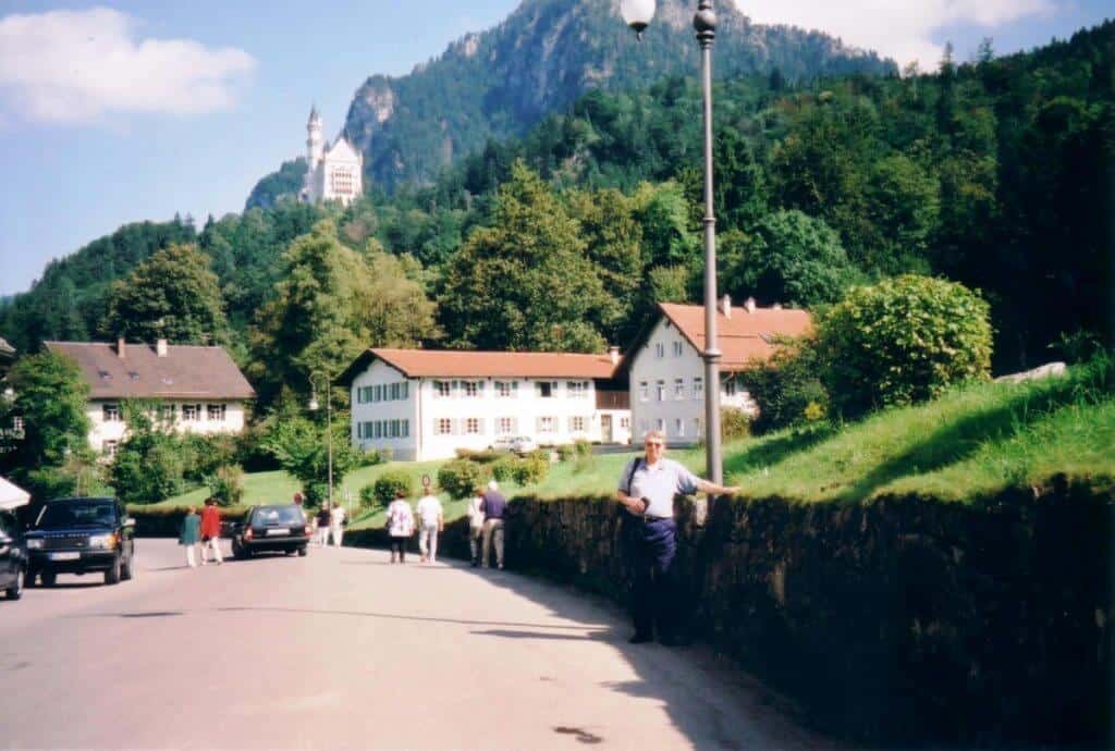 A serene street scene in Schwangau, Germany, with pedestrians and parked cars, a gentleman leaning against a wall, and the majestic Schloss Neuschwanstein Castle visible atop a lush green hill in the distance under a clear sky.