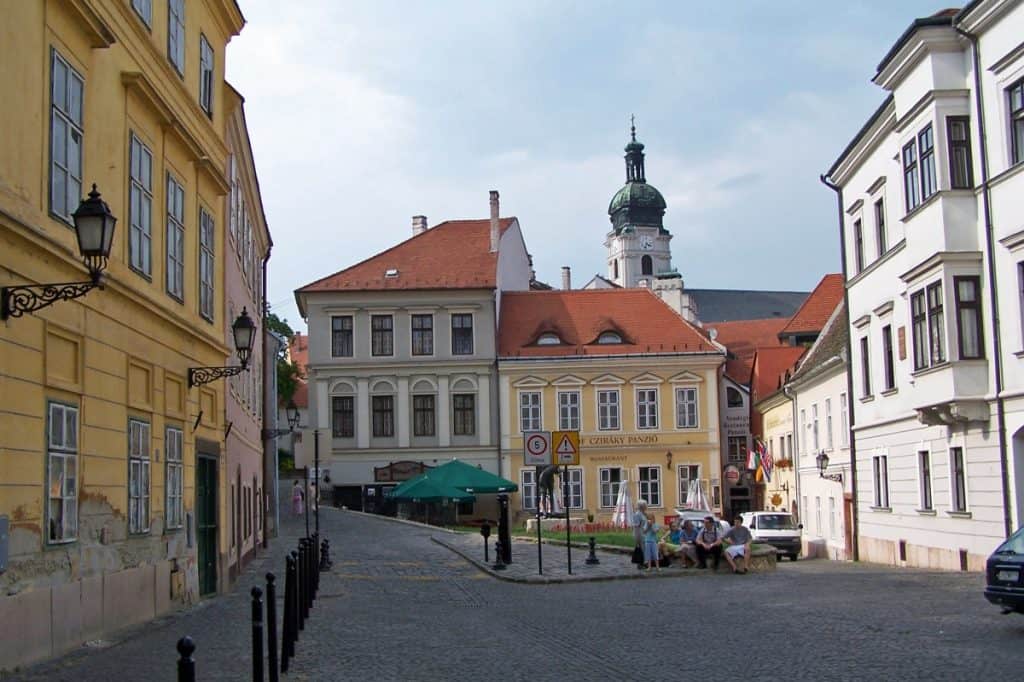 Charming cobblestone street in the historic town of Gyor, Hungary, flanked by traditional European buildings in pastel yellow and white. The street leads to a visible town square with a green and white umbrella cafe and a towering church with a green dome in the background, under a partly cloudy sky.