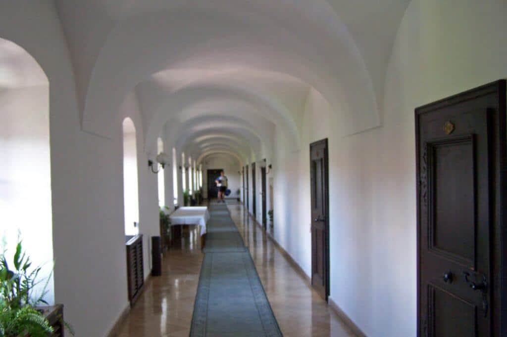 Long corridor of Hotel Klastrom in Gyor, showcasing its history as a former monastery with a series of classic arched ceilings and white walls. The hallway is lined with heavy wooden doors on one side and columns on the other, leading towards a distant figure, enhancing the serene and historic ambiance of the setting