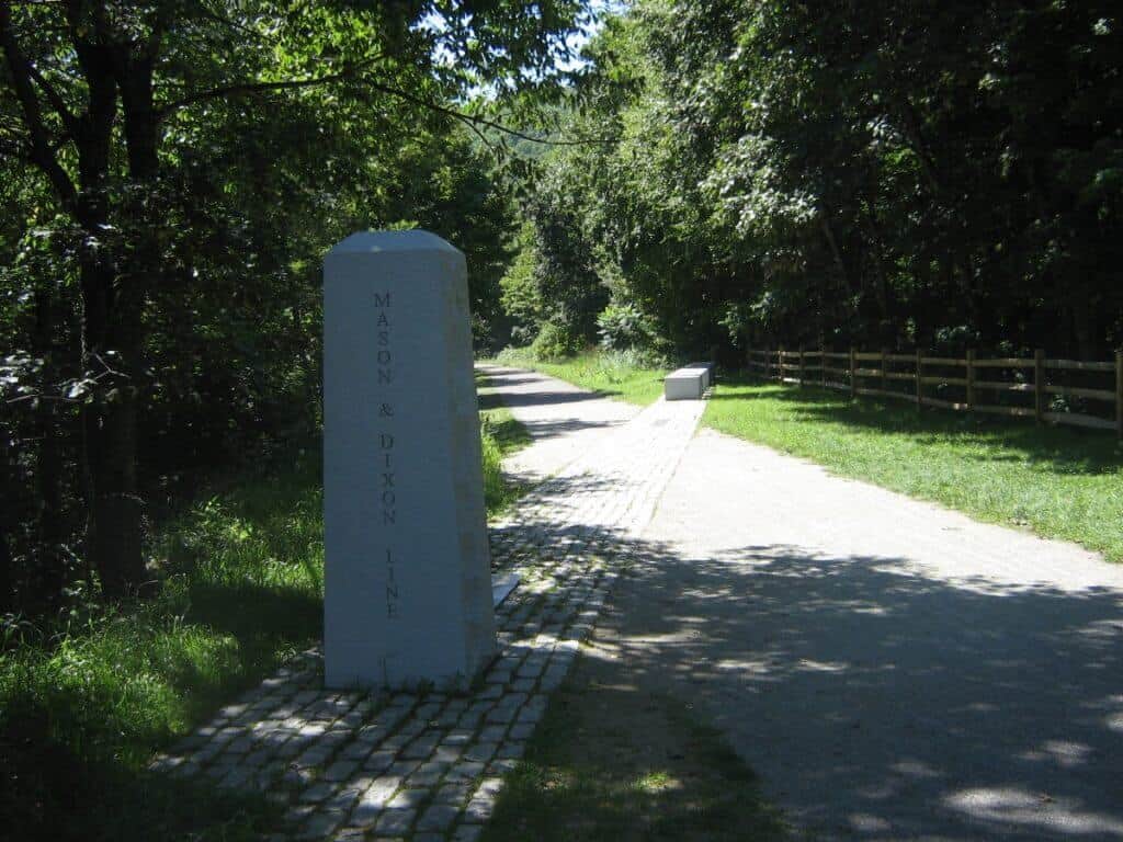 A historical marker for the Mason-Dixon Line beside the crushed limestone path of the Great Allegheny Passage Trail, surrounded by a tranquil setting of trees and wooden fencing.