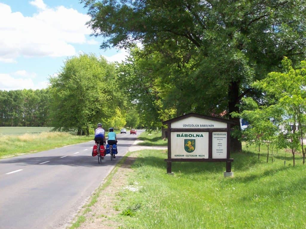 Two cyclists with pannier bags attached to their bikes ride along a road leading into Babolna, Hungary. A welcoming sign featuring the town's emblem is visible to the right, set against a backdrop of lush green trees and a clear blue sky