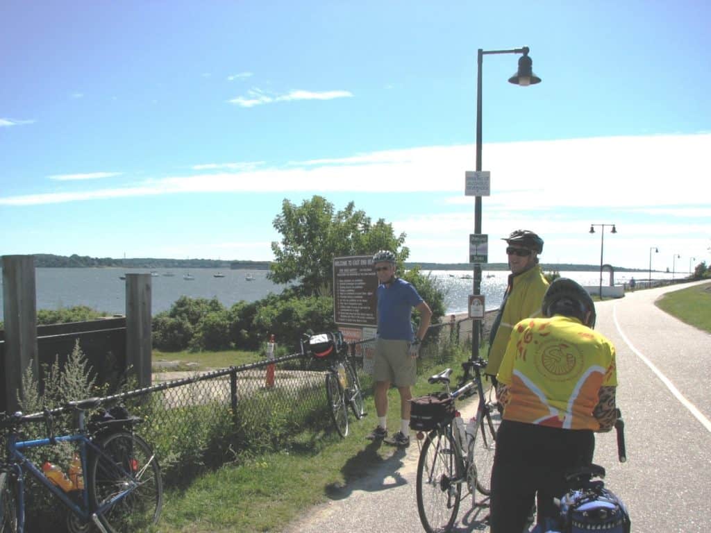Coastal Maine Cycling Routes with cyclists taking a break along the Eastern Promenade Trail in Portland, Maine, with their bikes parked by the trail, overlooking a scenic view of a calm bay dotted with boats under a blue sky,