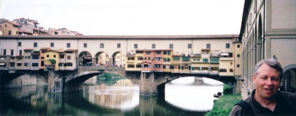 The iconic Ponte Vecchio bridge spans the Arno River in Florence, with its unique shops built along it, reflected in the water below. The architectural beauty of this medieval stone closed-spandrel segmental arch bridge is a historic symbol of Florence.