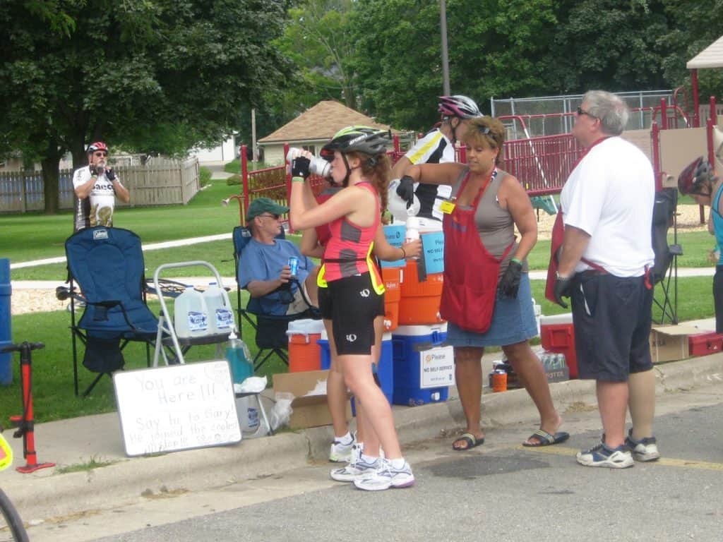 A rest stop during a Wisconsin bike tour in Brillion, featuring cyclists and volunteers. A female cyclist adjusts her helmet while talking to a volunteer in a red apron who is offering refreshments. Signs and coolers are set up along the roadside, and other participants are seen resting and hydrating. A man takes photos of the scene, capturing the community spirit and support at the event.
