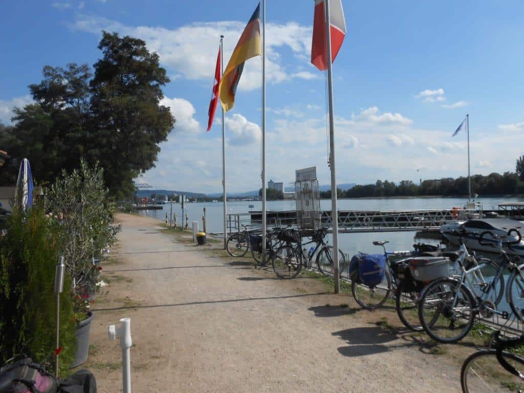 A docking area in Basel along the Rhine Cycling Route, with parked bicycles and fluttering flags of Switzerland, France, and Germany, representing the city's proximity to the tripoint of these nations.