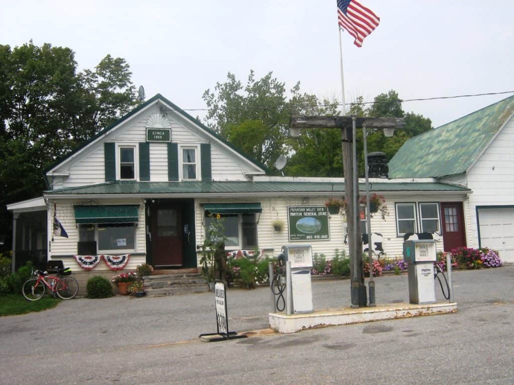 A quaint, historic general store at Panton Corner, Vermont, captured on a cloudy day. The store, marked as "Circa 1890" and "Panton Valley New Depot General Store," features a white facade adorned with American flags and floral decorations. In front, two vintage gas pumps stand, with a flagpole flying the American flag prominently beside them. A bicycle is parked on the porch, adding a charming, small-town feel to the scene.