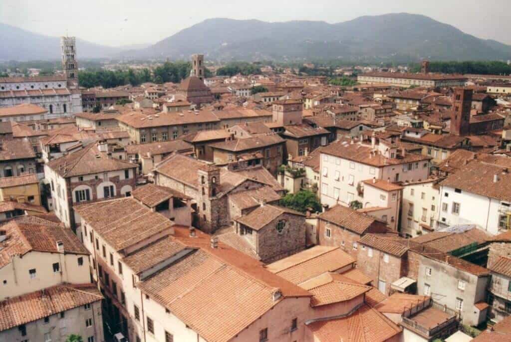 An aerial view of the dense, terracotta-roofed houses of Lucca, Tuscany, interwoven with narrow streets and dotted with several historic towers and church steeples, against the backdrop of the distant Tuscan hills.