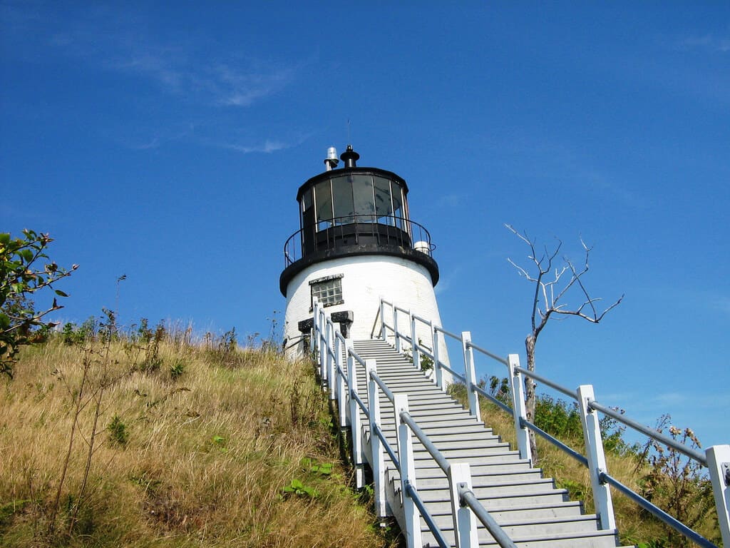 The Owls Head Lighthouse in Maine, standing atop a grassy slope with a clear blue sky overhead, is accessible via a white staircase flanked by a lone leafless tree.