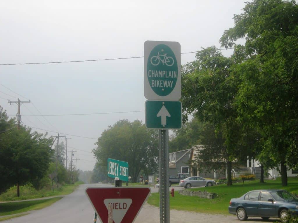 Directional sign for the Champlain Bikeway showing a green arrow pointing upwards, located at an intersection under overcast skies. The sign is positioned above a red 'YIELD' sign and a green 'JERSEY STREET' street sign. In the background, a residential street with houses and parked cars can be seen, shrouded in a light haze.