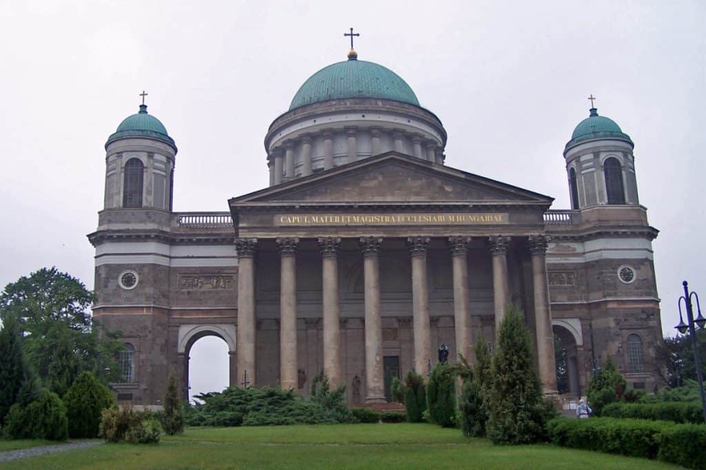 Front view of the imposing Esztergom Cathedral in Hungary, featuring a neoclassical architectural style with a massive dome and two prominent green-roofed bell towers. The cathedral's grand facade is adorned with columns and inscriptions, set against a backdrop of a cloudy sky, reflecting its historical and religious significance.