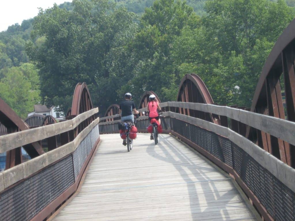 Two cyclists with fully loaded panniers biking side by side on a wooden and steel bridge along the Great Allegheny Passage, surrounded by dense green foliage