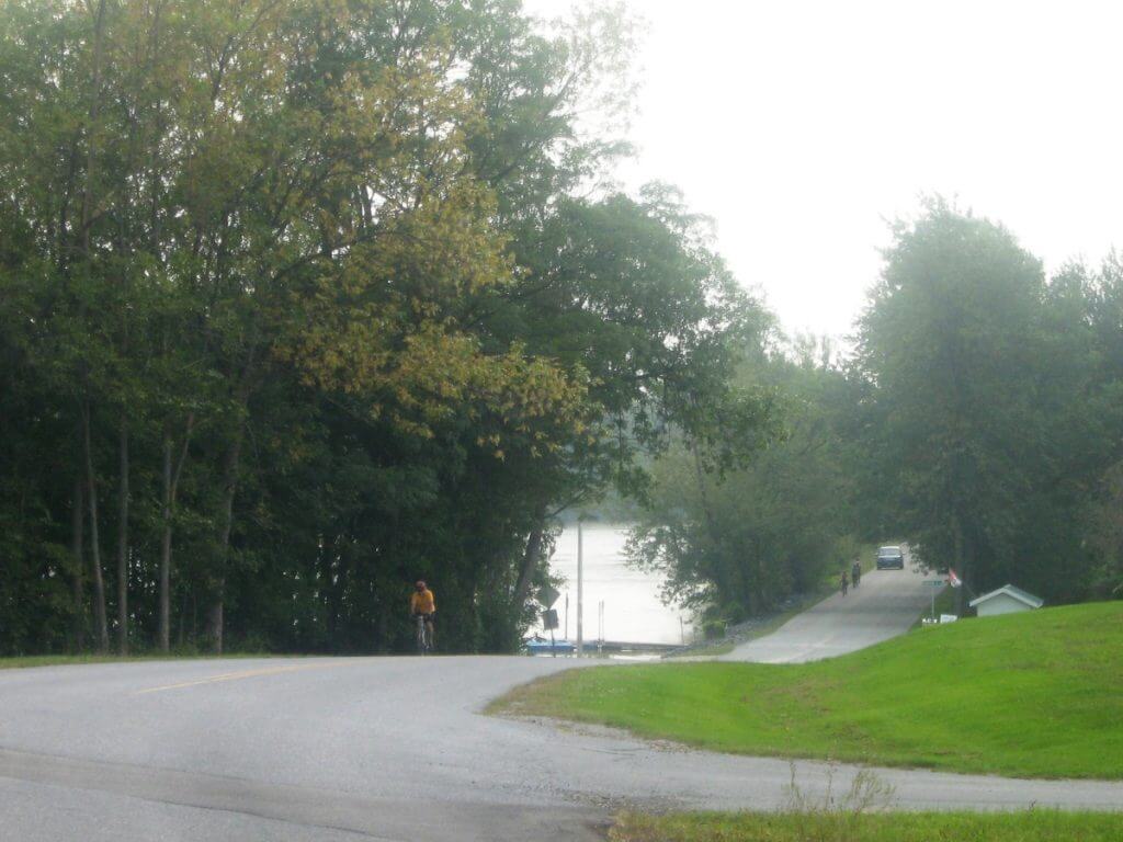 Misty morning on a curving road lined with lush green trees transitioning to autumn colors in Middlebury, Vermont. A cyclist in a yellow jacket is visible on Basin Harbor Road, heading towards a scenic lake visible at the road's end. The peaceful residential area features small houses and docks along the lakeside.
