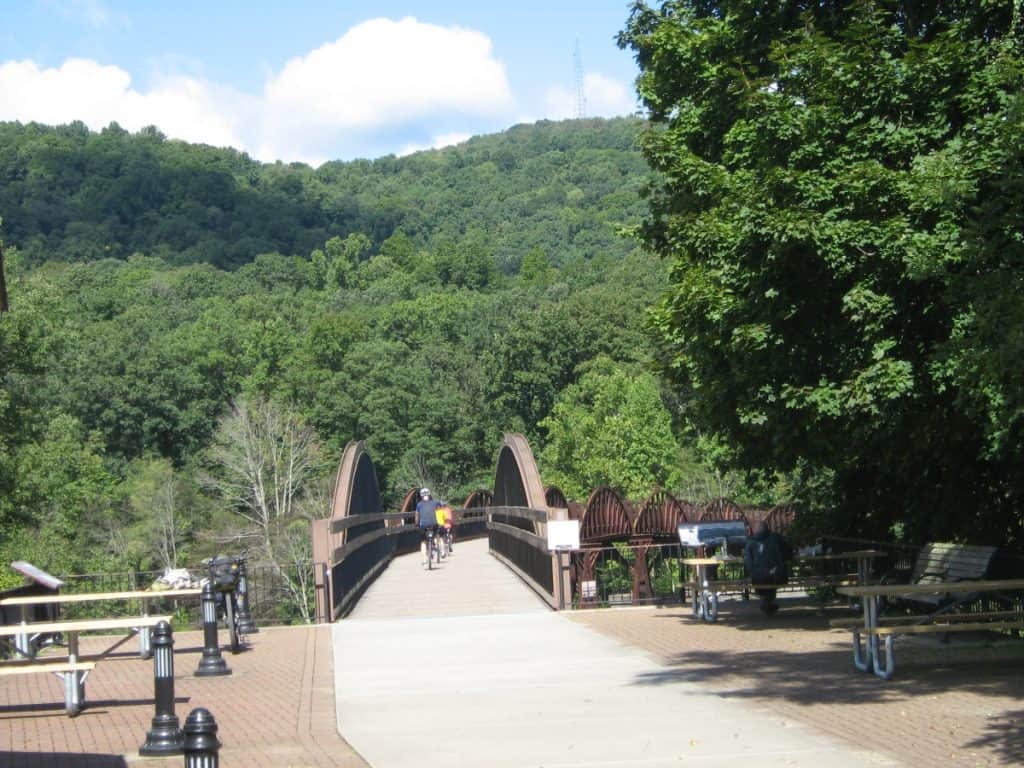 Two cyclists crossing a low-level bridge on the Great Allegheny Passage at Ohiopyle, with lush green trees on a hillside in the background under a clear blue sky