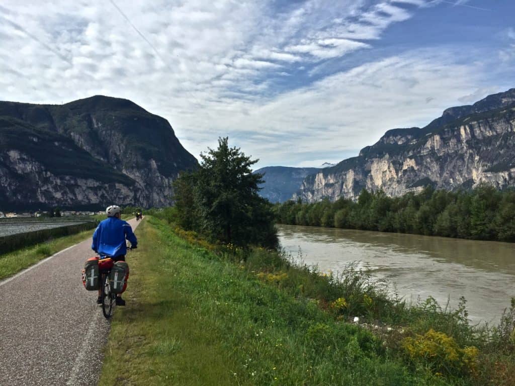 This image depicts a serene moment on the Adige cycle route between Bolzano and Trento, Italy. A cyclist, clad in a blue jacket and helmet, rides along a paved path that runs parallel to the river Adige. The path is bordered by lush greenery and wildflowers, with majestic mountains rising steeply on both sides.