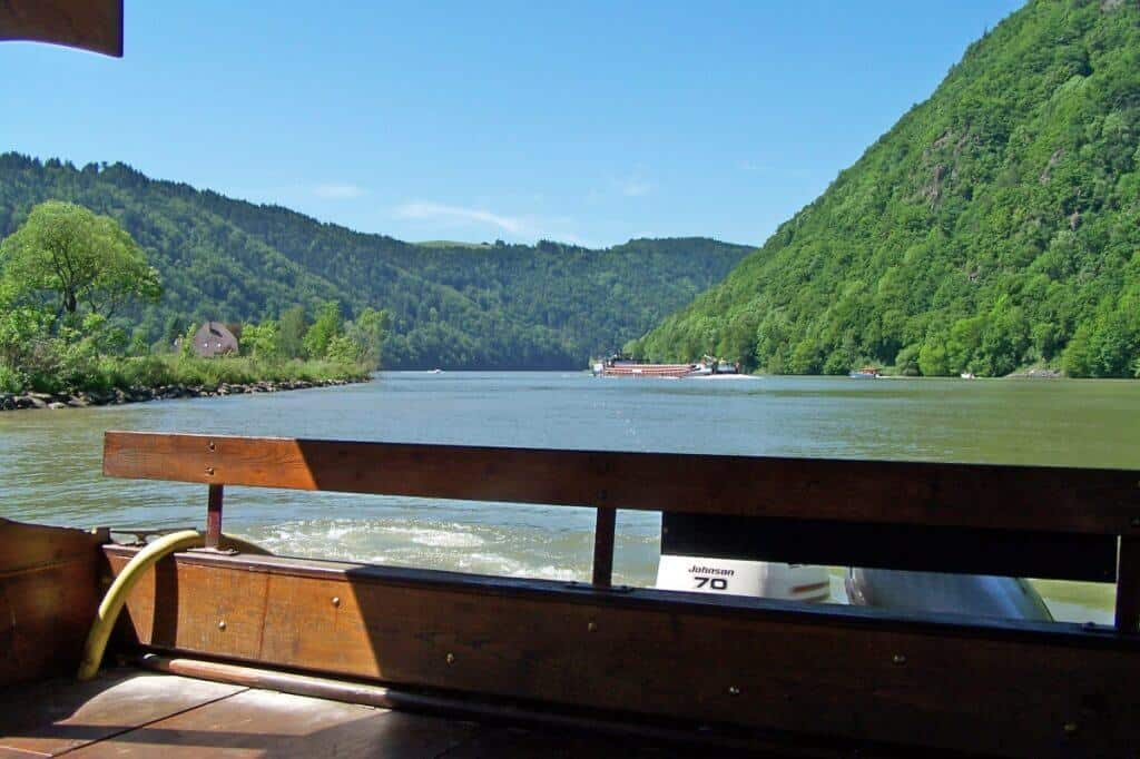 View from a bicycle ferry crossing the Schlögen Bend on the Danube River, showcasing the tranquil waters and verdant, forest-covered hills under a bright blue sky.