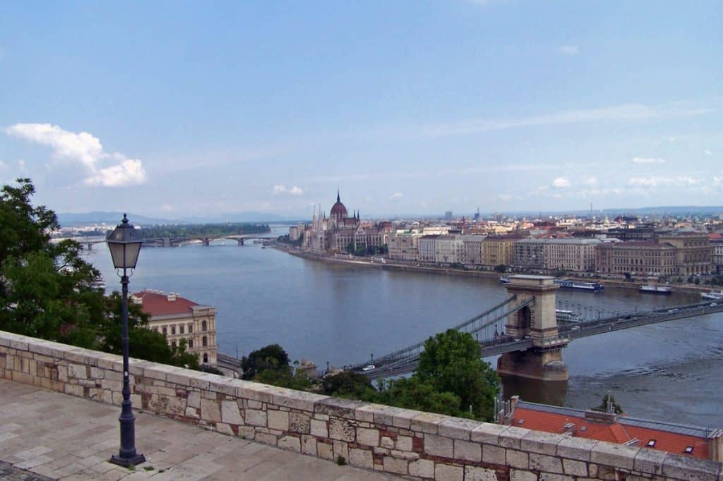 Panoramic view of Budapest from the Castle District, showing the Danube River flowing between the Buda and Pest sides of the city. The iconic Chain Bridge connects the two sides, with the Hungarian Parliament Building's distinctive Gothic architecture visible along the riverbank. The cityscape extends into the horizon under a bright sky with scattered clouds