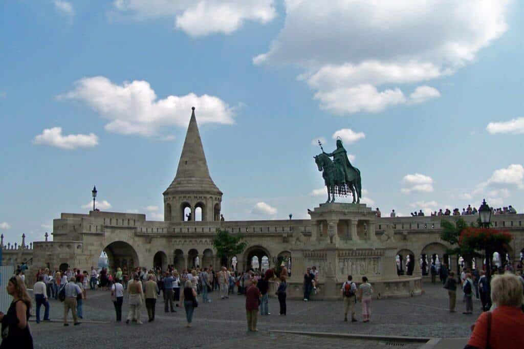 Busy scene at the Castle District in Budapest, featuring the iconic Fisherman's Bastion with its distinctively pointed towers and archways, and the equestrian statue of St. Stephen. Tourists gather around the historical structures, exploring and taking photos under a partly cloudy sky, contributing to a lively atmosphere at this famous Hungarian landmark