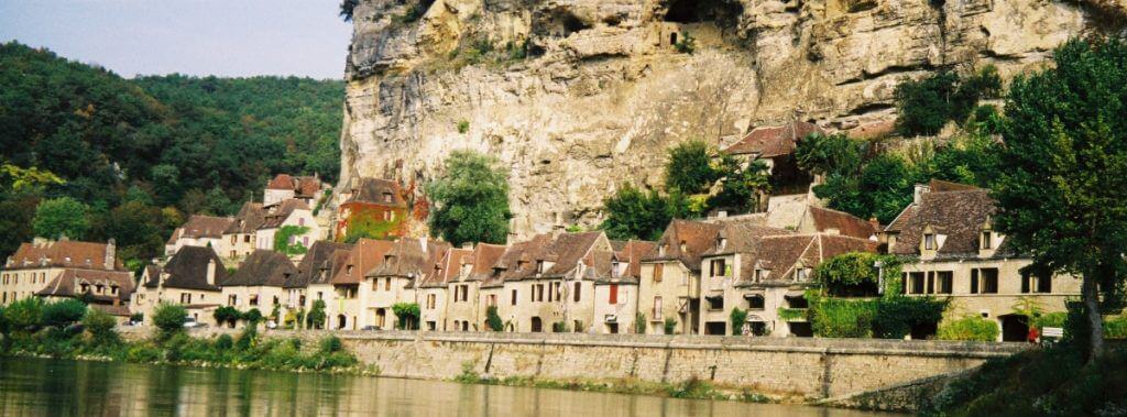 Panoramic view of La Roque-Gageac, France, with its beige traditional houses and terracotta roofs nestled against a limestone cliff beside the calm waters of the Dordogne River.