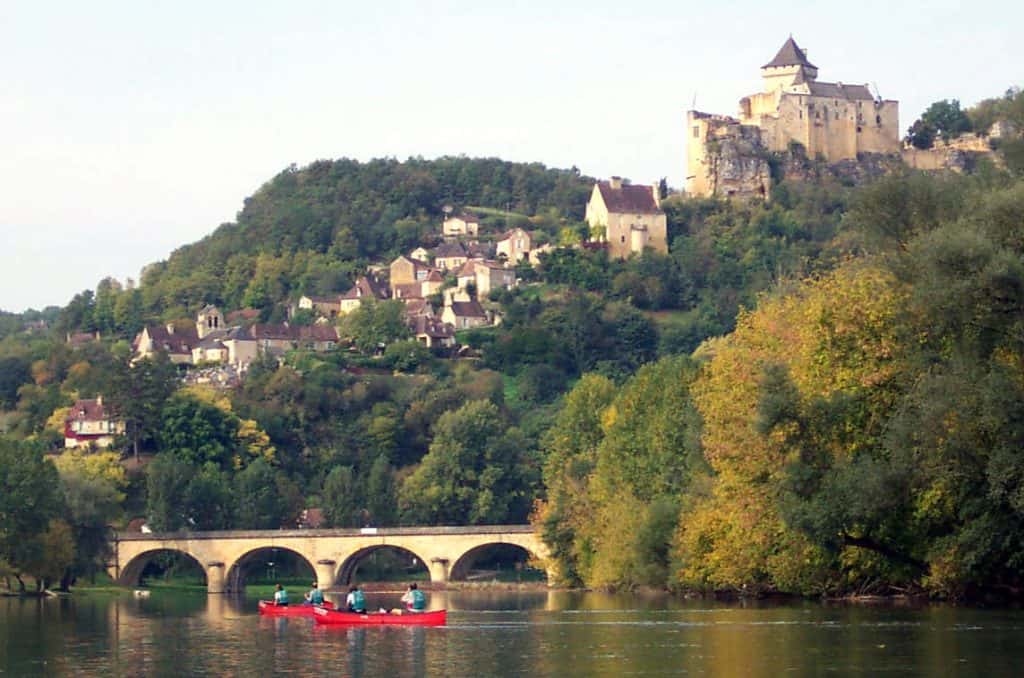 A tranquil scene on the Dordogne River in France with people canoeing in the foreground, and the historic Chรขteau de Castelnaud rising majestically on the hill above, framed by lush greenery and a stone bridge